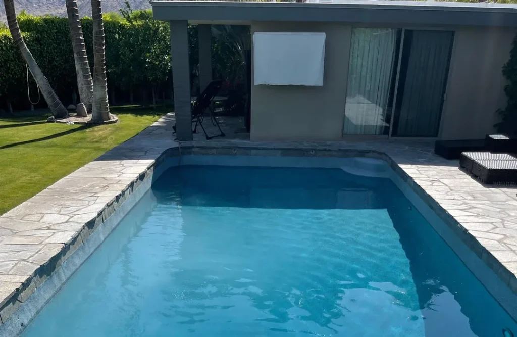 Rectangular swimming pool with blue water and stone coping, surrounded by a lawn and palm trees near a building.