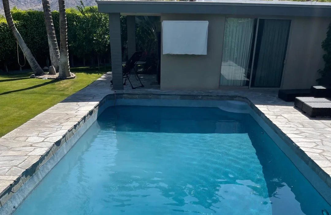 Rectangular swimming pool with blue water and stone coping, surrounded by a lawn and palm trees near a building.